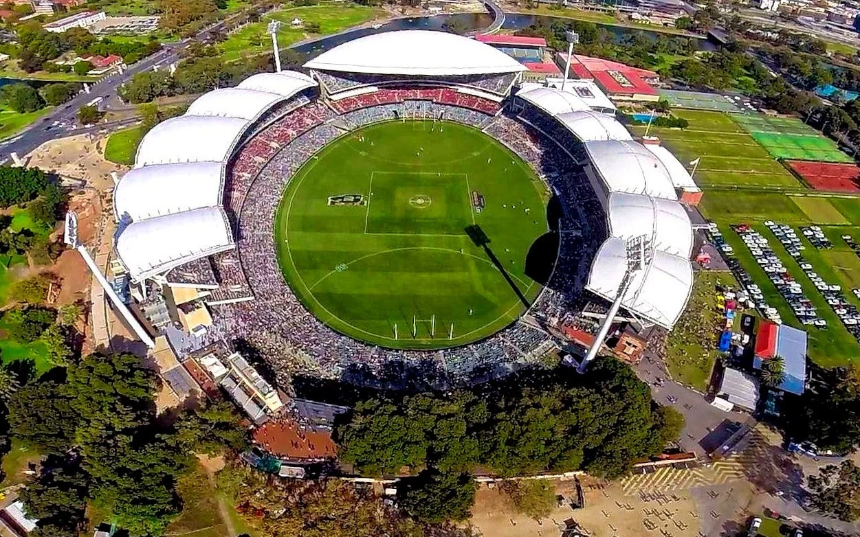Aerial wide view of Adelaide Oval with the oval field, stands and surrounding city greenery visible.